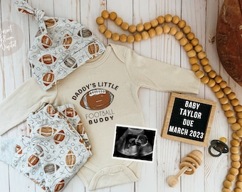 May include: A tan bodysuit with the text "Daddy's Little Football Buddy" and a football graphic. A gray and brown football-themed hat, a pair of gray and brown football-themed pants, a wooden rattle, a pacifier, a wooden letter board with the text "Baby Taylor Due March 2023", and a brown football are also pictured.