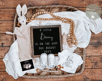 Peut inclure: Un tableau noir avec l'inscription "A little Bunny is on the way due August 2023". Un body blanc avec l'inscription "Baby Miller" est à côté du tableau noir. Une couverture blanche, un lapin en peluche, un hochet en bois, un biberon en bois et une paire de chaussures de bébé blanches sont également présents sur l'image.