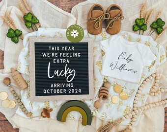 May include: A black letter board with the text "This year we're feeling extra lucky arriving October 2024".  A white onesie with the text "Baby Williams" is next to the letter board.  The onesie is on a beige blanket with gold and green accents.  There are also wooden beads, a wooden rattle, and a wooden toy.