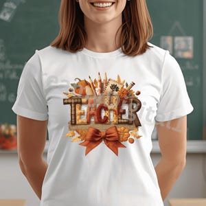 May include: White t-shirt with a fall-themed design. The word "TEACHER" is displayed in a decorative font, surrounded by pumpkins, leaves, pencils, and an orange bow. The shirt is a short-sleeved crew neck.