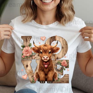 以下が含まれることがあります： 茶色のハイランド牛のイラストと木製の文字で「LOVE」という文字が描かれた白いTシャツ。デザインにはピンクのバラとハートが含まれており、素朴で魅力的な美学を作り出しています。