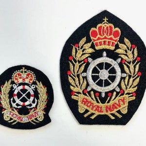 Op de afbeelding: Twee geborduurde patches met een zwarte achtergrond, gouden rand en rode accenten. De grotere patch heeft een gouden kroon, een zilveren stuurwiel en de tekst "ROYAL NAVY". De kleinere patch heeft een gouden kroon, twee zilveren ankers en een reddingsboei.