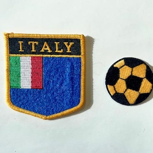 Puede incluir: Dos parches bordados: uno con forma de escudo con la palabra "ITALIA" y la bandera italiana, y un balón de fútbol. El escudo tiene un fondo azul y un ribete dorado. El balón es negro y dorado.