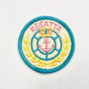 May include: A white embroidered patch with a blue circle around the edge. The circle has a yellow wreath with a pink anchor and a crown inside. The text "REGATTA" is embroidered in pink around the top of the circle.