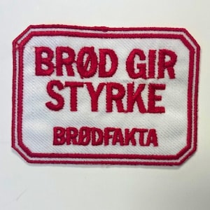 May include: Red embroidered patch with a white background. The patch says "Brod Gir Styrke" and "BrodFakta".