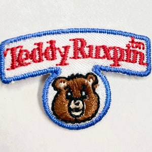 May include: A white patch with blue edging featuring the text "Teddy Ruxpin" in red lettering. Below the text is a brown embroidered bear face with a blue circle around it.