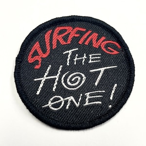 May include: Black round patch with white stitching. The text "SURFING THE HOT ONE!" is embroidered in white with a red "SURFING" and a swirl design in the "HOT".