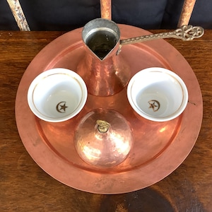 May include: A copper coffee set on a round tray. The set includes a copper coffee pot with a decorative handle, two small white cups with gold rims and a star and crescent design, and a copper lid.
