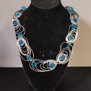 Può includere: Una collana in filo argentato con una serie di forme ovali e a spirale interconnesse. Ogni forma contiene una perla quadrata, traslucida, azzurra. La collana è esposta su un busto di gioielli nero.