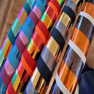 Könnte beinhalten: Eine Sammlung von Hula-Hoop-Reifen, die in einer Reihe angeordnet sind und jeweils ein eigenes Farbschema aufweisen. Die Reifen sind mit abwechselnden Bändern aus metallischem, irisierendem und mattem Klebeband in Orange, Rot, Blau, Grün, Gold und Schwarz umwickelt.