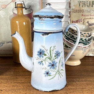 May include: A blue and white enamel coffee pot with a floral design. The pot has a spout and a handle. The pot is sitting on a wooden table. There is a sign in the background that says "Comestibles aux Truffes".