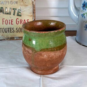 Peut inclure: Un petit pot rond en terre cuite avec un glaçage vert et marron. Une cafetière vintage en émail bleu et blanc avec des motifs floraux est en arrière-plan. Une enseigne ancienne est également visible.