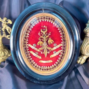 Peut inclure: Un reliquaire religieux ovale encadré avec un fond en velours rouge, des ornements dorés et de petites étiquettes blanches. Deux chandeliers dorés et un crucifix doré sont en arrière-plan. Le cadre est bleu foncé.