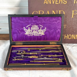 Peut inclure: Un microscope en laiton antique et une boîte en bois contenant des instruments de dessin. La boîte, ouverte, révèle des instruments en laiton sur un intérieur en velours violet. Un livre avec l'inscription dorée "Exposition Internationale Anvers 1894 Grand Prix" est en arrière-plan.