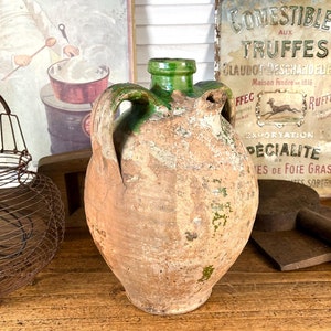 Peut inclure: Un pichet en terre cuite rustique avec un dessus et des anses en émail vert. Le pichet est posé sur une table en bois avec un panier en fil de fer, une enseigne vintage annonçant des truffes et une planche à découper en bois.