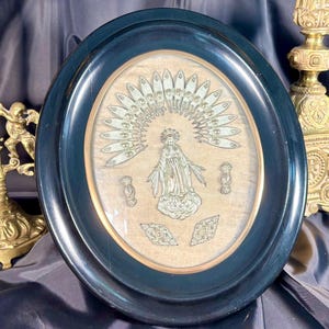 Puede incluir: Imagen religiosa enmarcada ovalada con marco azul oscuro. La imagen presenta una ilustración detallada de una figura religiosa con halo y detalles ornamentados. Un crucifijo y un candelabro dorados están en el fondo.