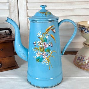 May include: A vintage, pale blue enamel coffee pot with a floral pattern. The pot has a long spout, a handle, and a lid with a small knob. The floral design features white, purple, and yellow flowers. The pot is displayed with other vintage items.