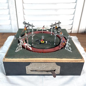 Puede incluir: Juego de carreras de caballos mecánico vintage con base negra y superficie de juego de fieltro verde. El juego presenta una pista circular con caballos y jinetes en miniatura. El texto "Jeu de Course" es visible en la parte delantera.