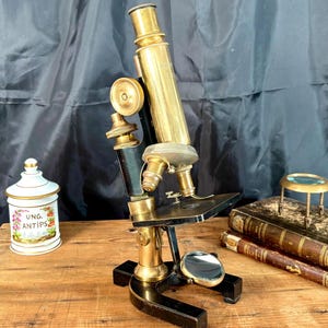 Peut inclure: Un microscope en laiton et métal noir avec une platine circulaire, des boutons de réglage et un miroir réfléchissant. À côté se trouvent un pot en céramique blanc étiqueté Ung. Antips, deux livres reliés en cuir et une loupe en laiton.