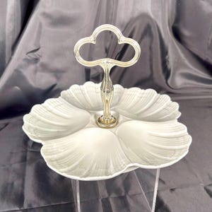 Peut inclure: Un plat de service blanc à plusieurs niveaux avec une poignée argentée. Le plat a un design festonné en forme de pétale et est soutenu par une tige centrale ornée. La poignée a la forme d'une fleur stylisée.