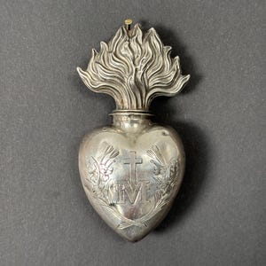 May include: A silver-toned metal heart-shaped ex-voto ornament featuring a flame motif at the top. The center of the heart is engraved with a cross above the letter M, flanked by floral engravings on both sides. A small metal pin protrudes from the top.