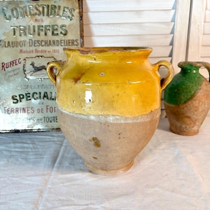 Puede incluir: Una olla de cerámica grande, amarilla y marrón con dos asas, de aspecto rústico. A la derecha hay una jarra más pequeña, verde y marrón. Un letrero de metal vintage con texto en francés está en el fondo.