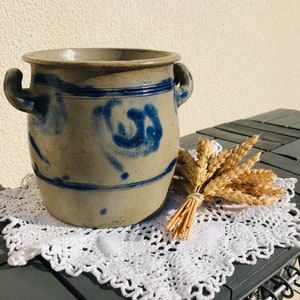 Könnte beinhalten: Ein antiker Steinzeugtopf mit blau-weißen Bemalungen. Der Topf hat zwei Henkel und steht auf einer weißen Häkeldeckchen.