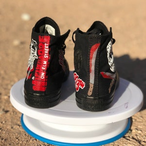 May include: A pair of black high-top Converse sneakers with a custom design featuring a white and red Freddy Krueger character from the movie "A Nightmare on Elm Street". The sneakers have the text "A Nightmare On Elm Street" on the side.