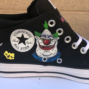 May include: Black Converse Chuck Taylor All Star low-top trainers with a hand-painted design featuring a cartoon clown with green hair, a red nose, and a blue collar. The Converse logo is visible on the side of the shoe.