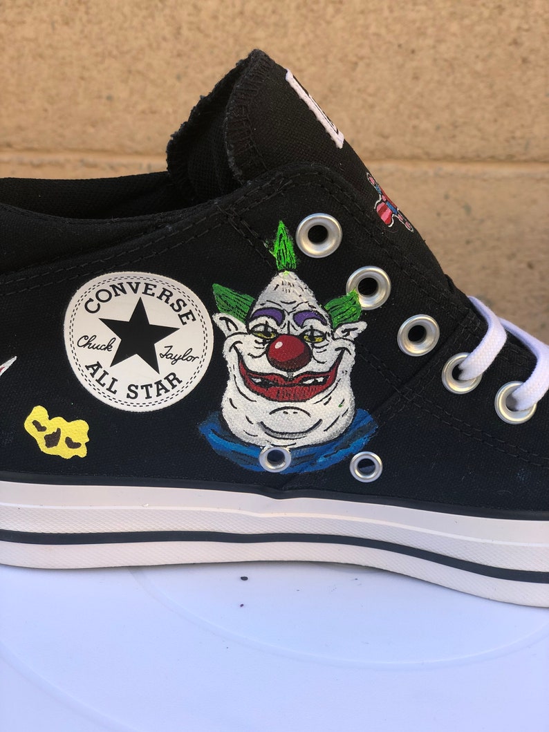 May include: Black Converse Chuck Taylor All Star low-top trainers with a hand-painted design featuring a cartoon clown with green hair, a red nose, and a blue collar. The Converse logo is visible on the side of the shoe.