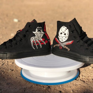 May include: A pair of black high-top trainers with custom painted designs. One shoe features Freddy Krueger from the Nightmare on Elm Street film franchise, and the other shoe features Jason Voorhees from the Friday the 13th film franchise.