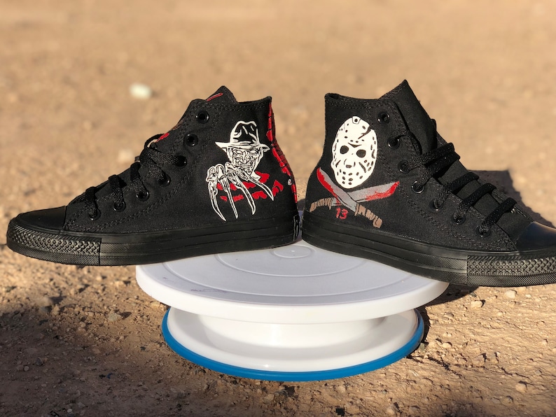 May include: A pair of black high-top trainers with custom painted designs. One shoe features Freddy Krueger from the Nightmare on Elm Street film franchise, and the other shoe features Jason Voorhees from the Friday the 13th film franchise.