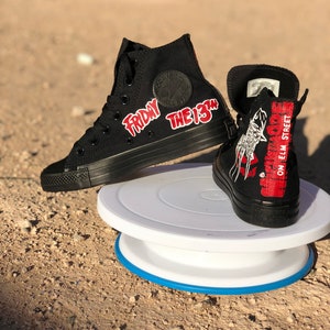 May include: A pair of black high-top Converse trainers with custom designs. One shoe features the text "Friday the 13th" in red lettering. The other shoe features a graphic of a hockey mask and the text "Nightmare on Elm Street".