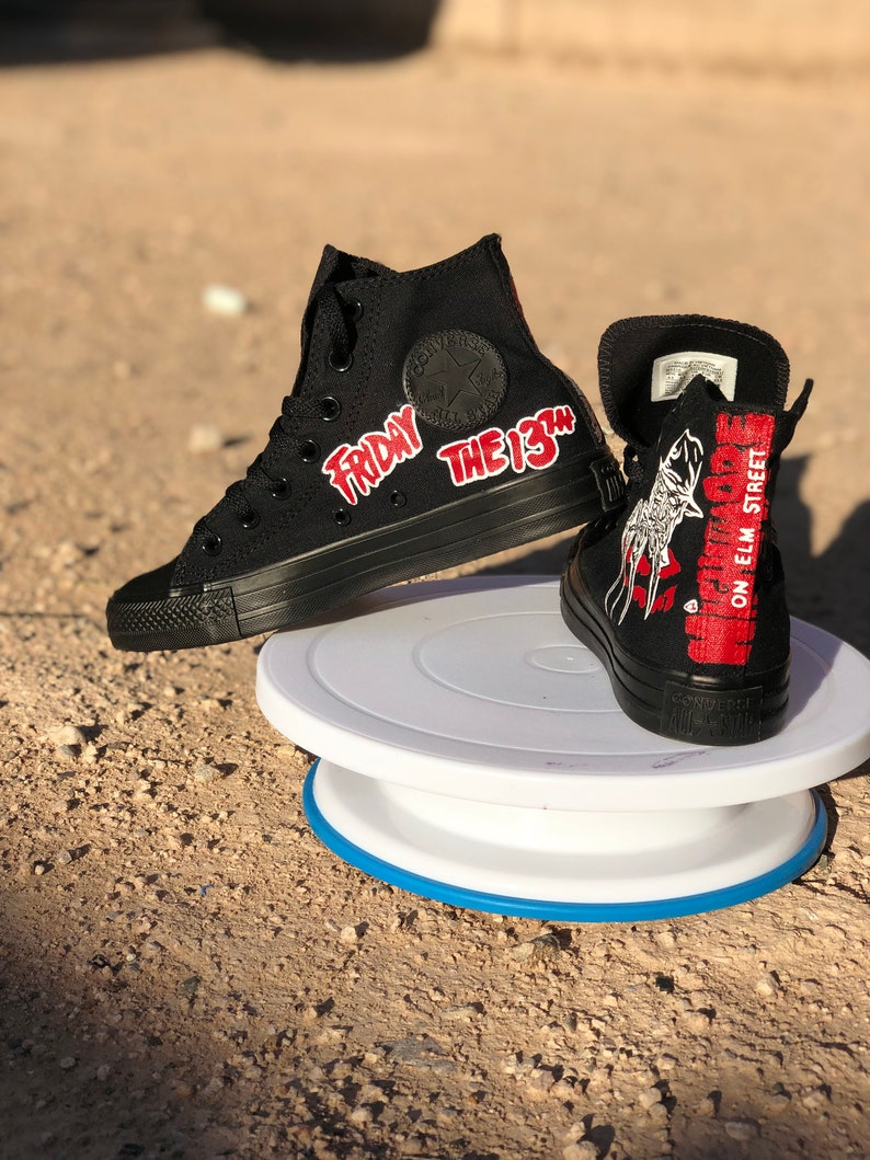 May include: A pair of black high-top Converse trainers with custom designs. One shoe features the text "Friday the 13th" in red lettering. The other shoe features a graphic of a hockey mask and the text "Nightmare on Elm Street".