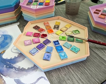 May include: A hexagonal watercolor palette with a rainbow of colors in individual pans. The palette is made of plastic and has a light blue, pink, and beige color scheme. A paintbrush is resting on the palette.