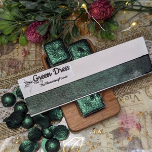 May include: Green Dress watercolor paint by The Blooming Faerie. The paint is a deep green with a metallic sheen. The paint is shown in a pan and in a swatch.