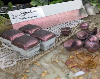 Pode incluir: Três pequenos quadrados de tinta rosa dourada metálica com base branca. A tinta é etiquetada como "Mojave Eclipse" e "Paint by The Blooming Faerie".