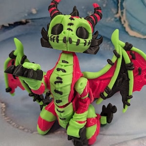 May include: A 3D-printed dragon figurine in green, red, and black. The dragon has large wings, horns, and a stitched-mouth design. The dragon is posed with its wings spread and its arms out.
