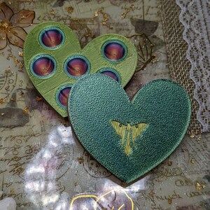 May include: Two green heart-shaped resin coasters with a textured surface. One coaster features a butterfly design and the other has a circular pattern with a red and purple centre.