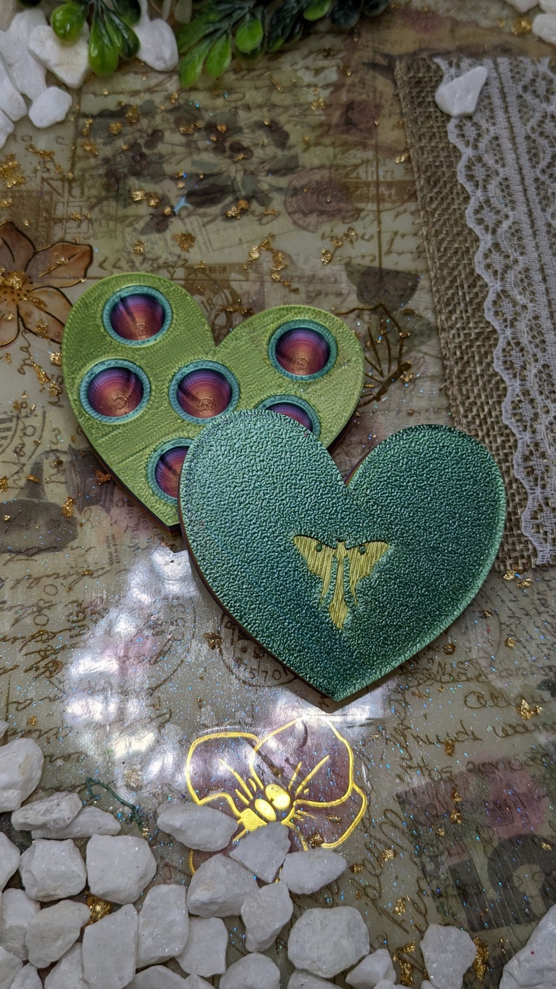 May include: Two green heart-shaped resin coasters with a textured surface. One coaster features a butterfly design and the other has a circular pattern with a red and purple centre.