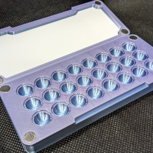 May include: A purple plastic case with a hinged lid and a grid of 25 small, round, indented compartments. The case is designed to hold small items, such as pills or beads.
