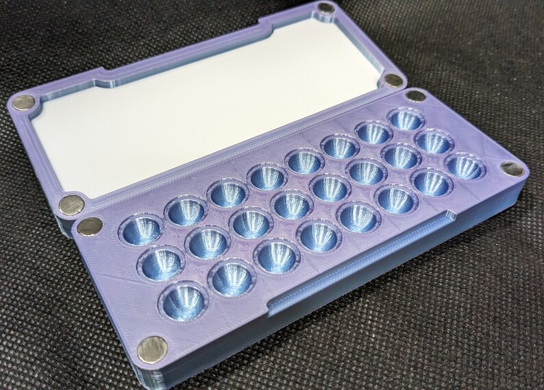 May include: A purple plastic case with a hinged lid and a grid of 25 small, round, indented compartments. The case is designed to hold small items, such as pills or beads.