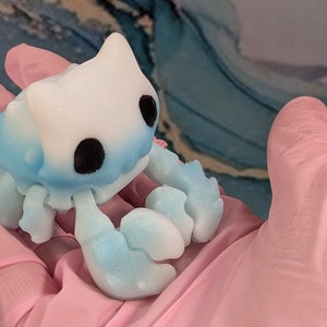 Pode incluir: Uma pequena figura de caranguejo de cer&acirc;mica pintada &agrave; m&atilde;o, azul e branca, com olhos pretos. O caranguejo est&aacute; sentado em uma superf&iacute;cie rosa.