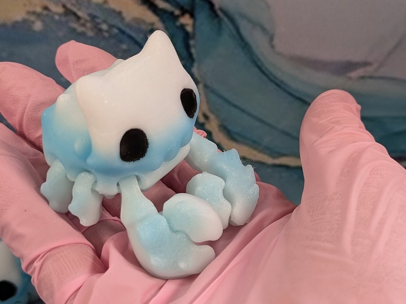 Pode incluir: Uma pequena figura de caranguejo de cer&acirc;mica pintada &agrave; m&atilde;o, azul e branca, com olhos pretos. O caranguejo est&aacute; sentado em uma superf&iacute;cie rosa.