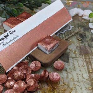 May include: A copper-colored watercolor paint pan with the name "Engulfed Rose" and the text "Paint by The Blooming Faerie". The pan is on a wooden block with a copper-colored paint swatch next to it. There are several copper-colored paint chips scattered around the pan.