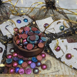 May include: A bowl filled with iridescent, multi-colored, round, resin earrings. The earrings are displayed on white cards with gold stars and moons. The earrings are packaged in a black mesh bag with gold stars.