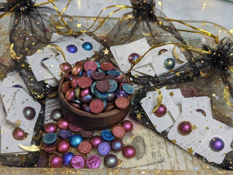 Pu&ograve; includere: Una ciotola piena di orecchini rotondi in resina iridescente e multicolore. Gli orecchini sono esposti su cartoncini bianchi con stelle e lune dorate. Gli orecchini sono confezionati in una borsa di rete nera con stelle dorate.