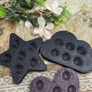 May include: Three silicone fidget toys in the shapes of a star, cloud, and heart. The star and cloud are black with a speckled texture, and the heart is purple with a speckled texture.