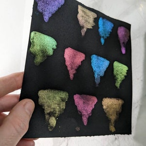 Pu&ograve; includere: Un foglio di carta nero con 12 campioni triangolari di vernice iridescente in vari colori, tra cui verde, blu, viola e oro.
