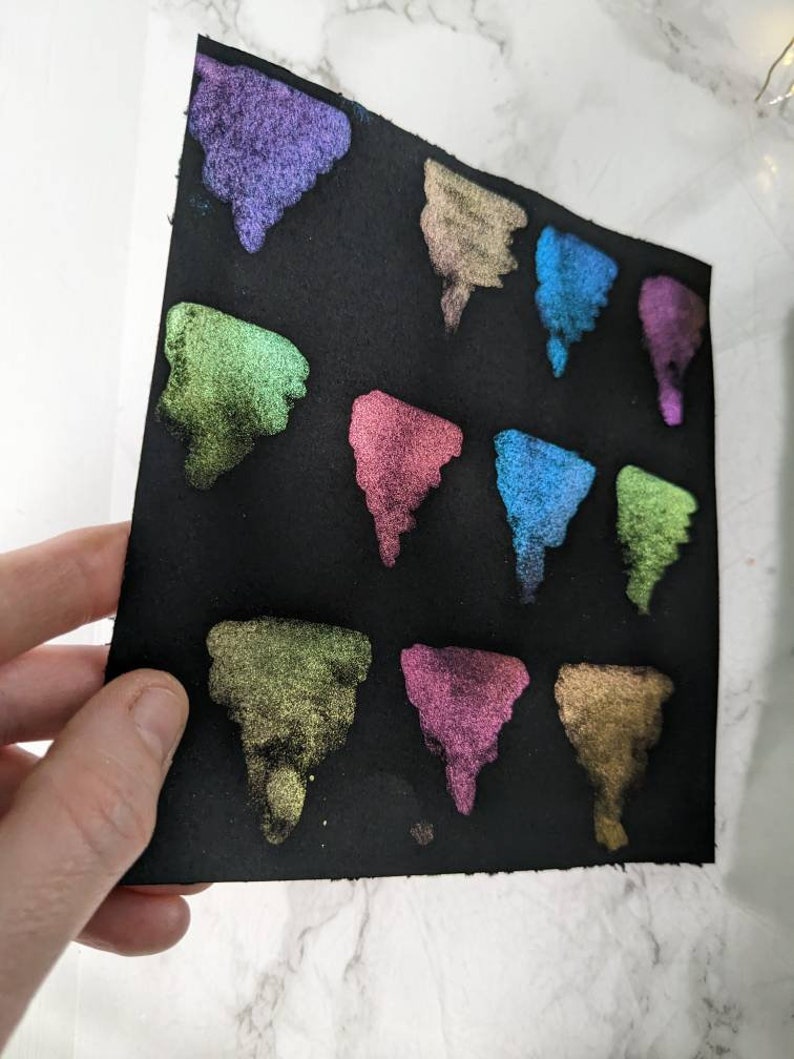 Pu&ograve; includere: Un foglio di carta nero con 12 campioni triangolari di vernice iridescente in vari colori, tra cui verde, blu, viola e oro.